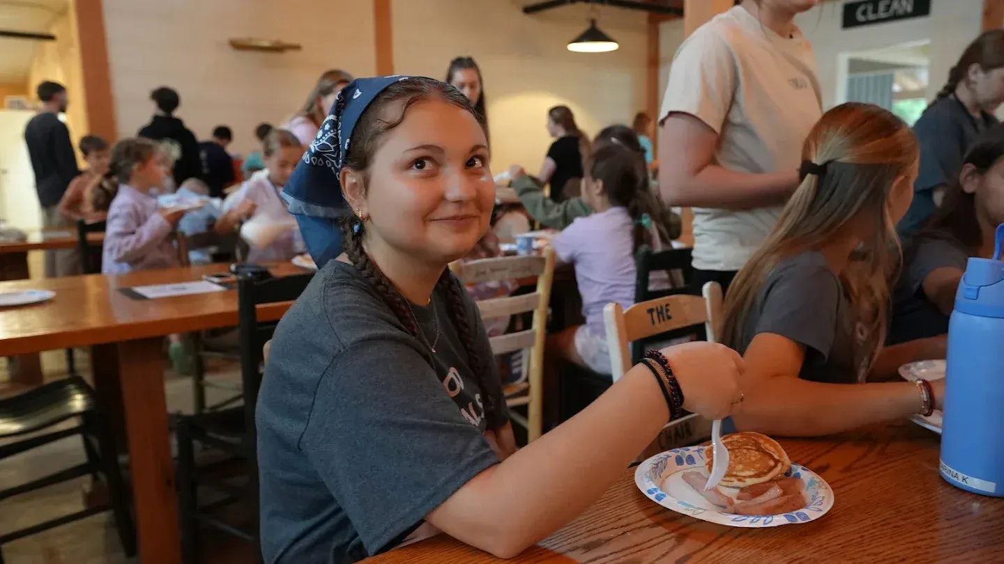 A camper in a team bandana smiles over pancakes in the dining hall.